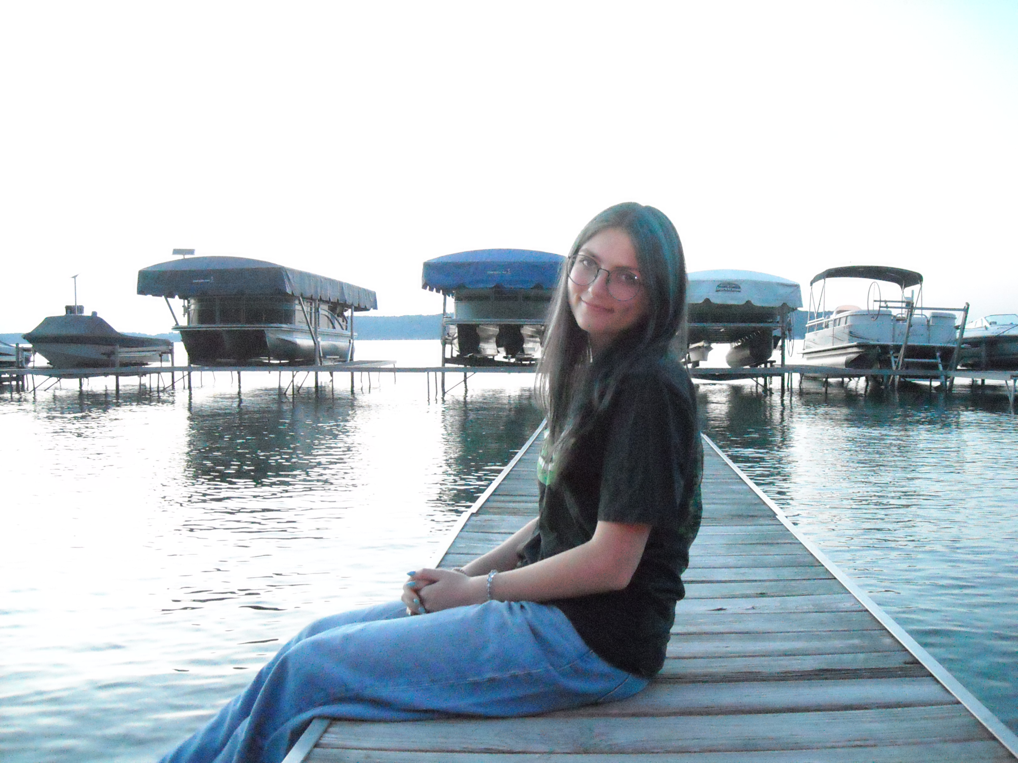 A girl sitting on a dock in front of some boats docked on a lake.