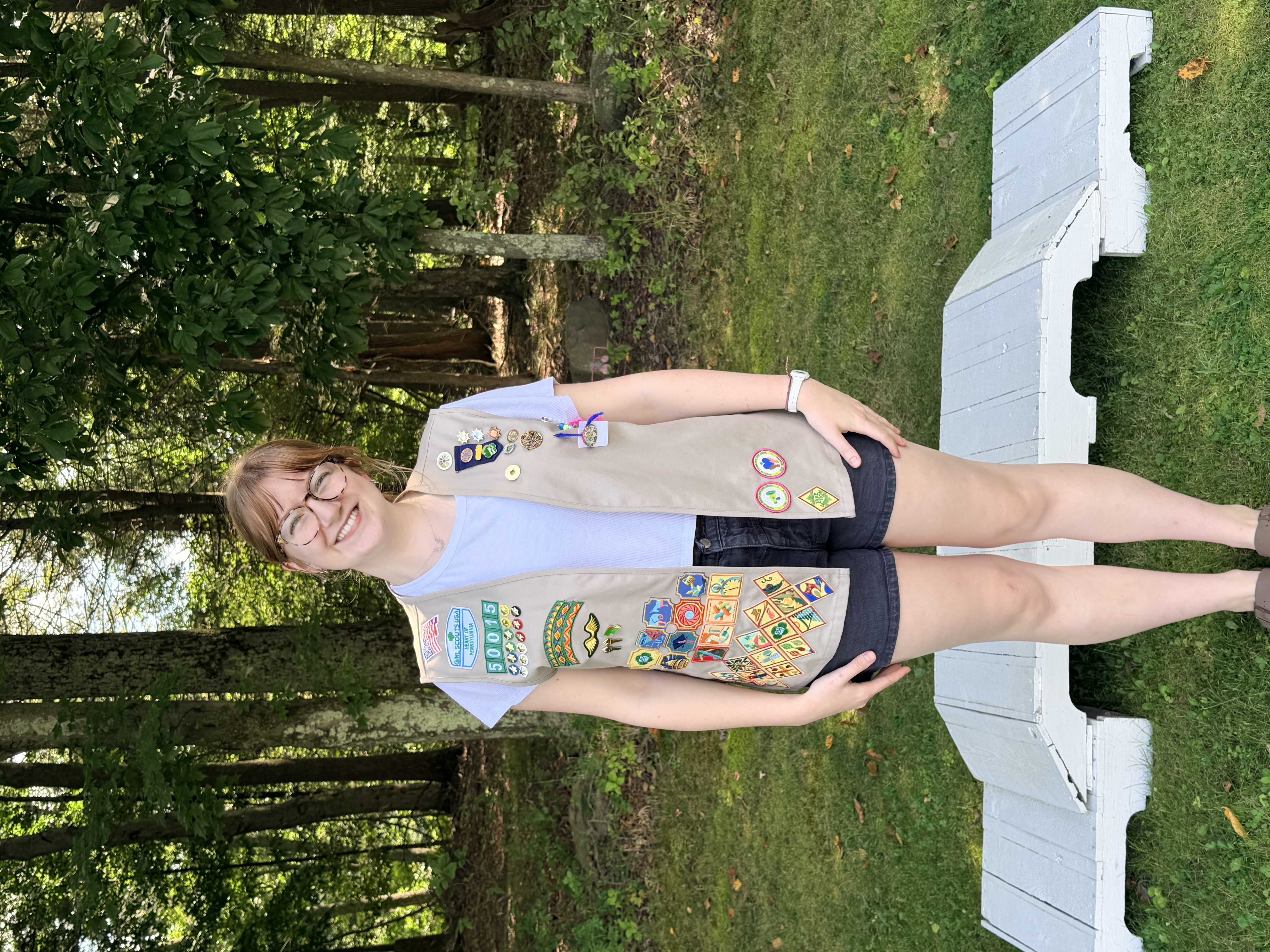 A picture of a young woman smiling in front of a small white bridge. The girl and the bridge are in front of a forrest. The girl is wearing a girl scout vest.